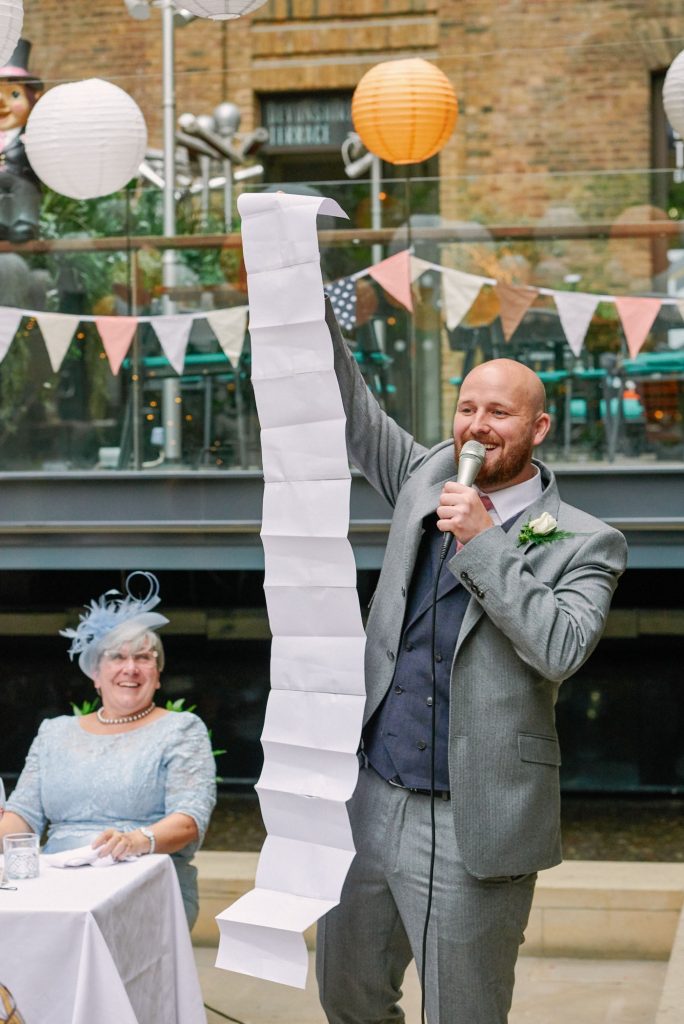 Best man laughing and holding up a long piece of paper during his wedding speech at Devonshire Terrance in Central London.
