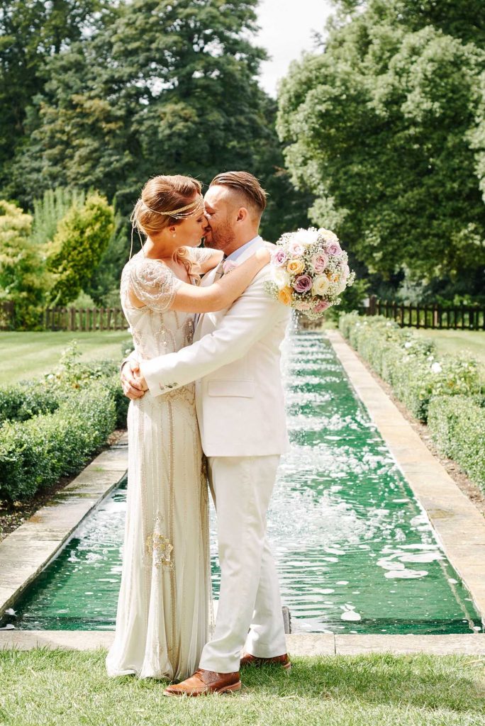 Bride and Groom kissing in front of a water feature whilst holding flowers on a sunny day at Woodhall Manor