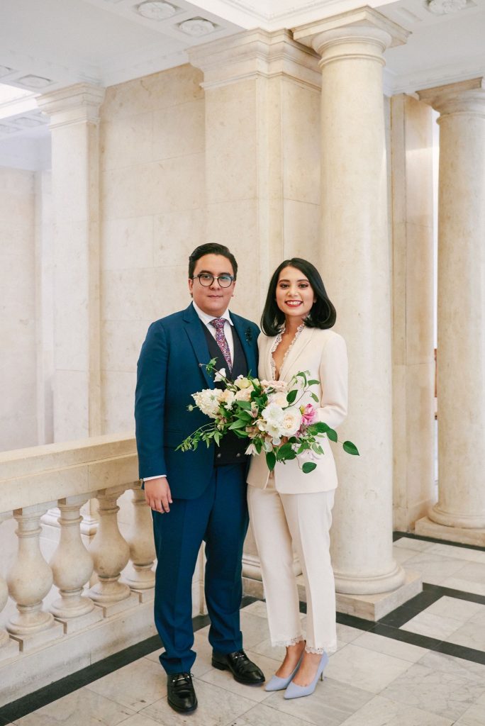 bride wearing white pantsuit with groom n blue suit at Old Marylebone Town Hall in Central London.