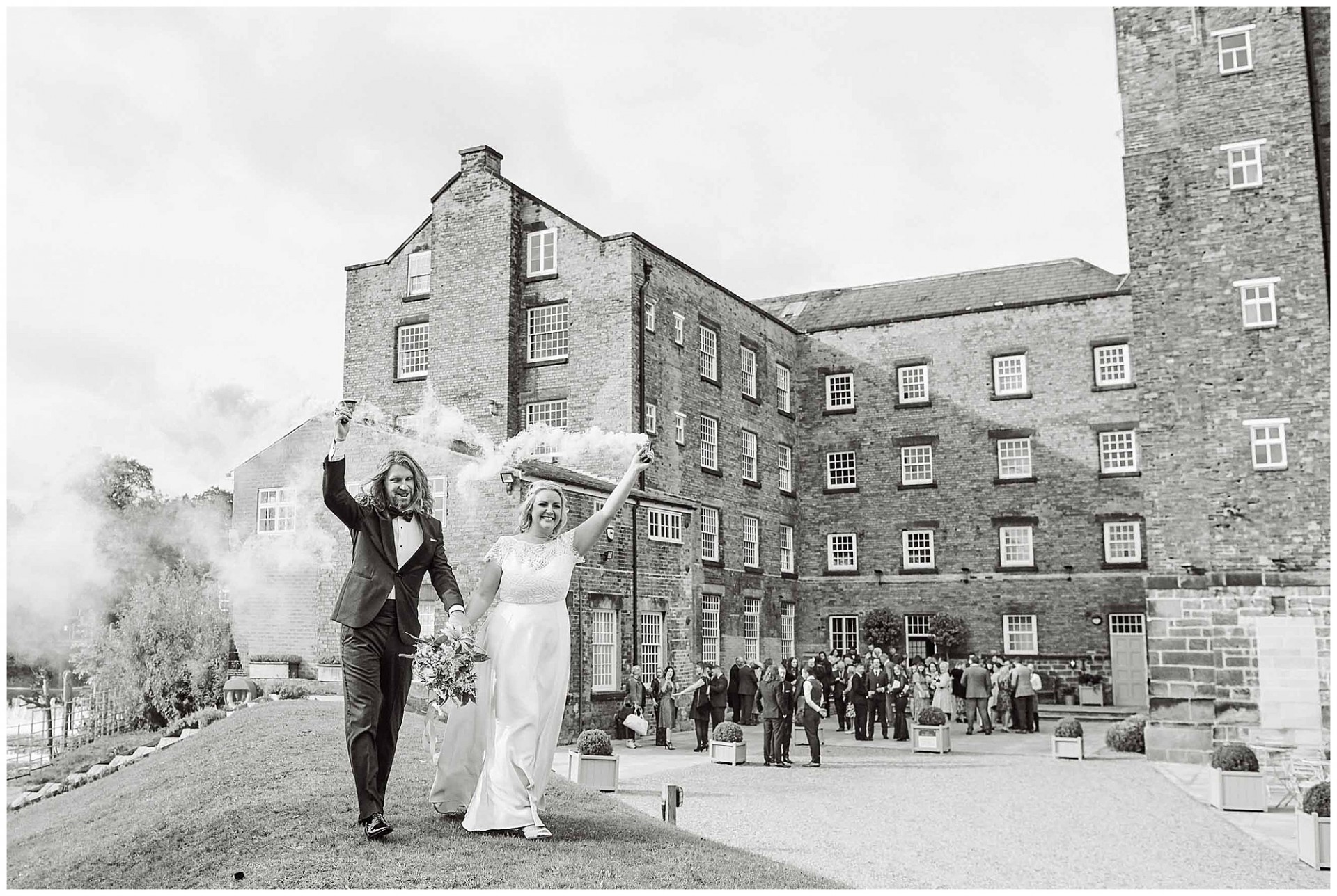 Bride and groom holding smoke bombs with the industrial West Mill Wedding Venue in the background. 