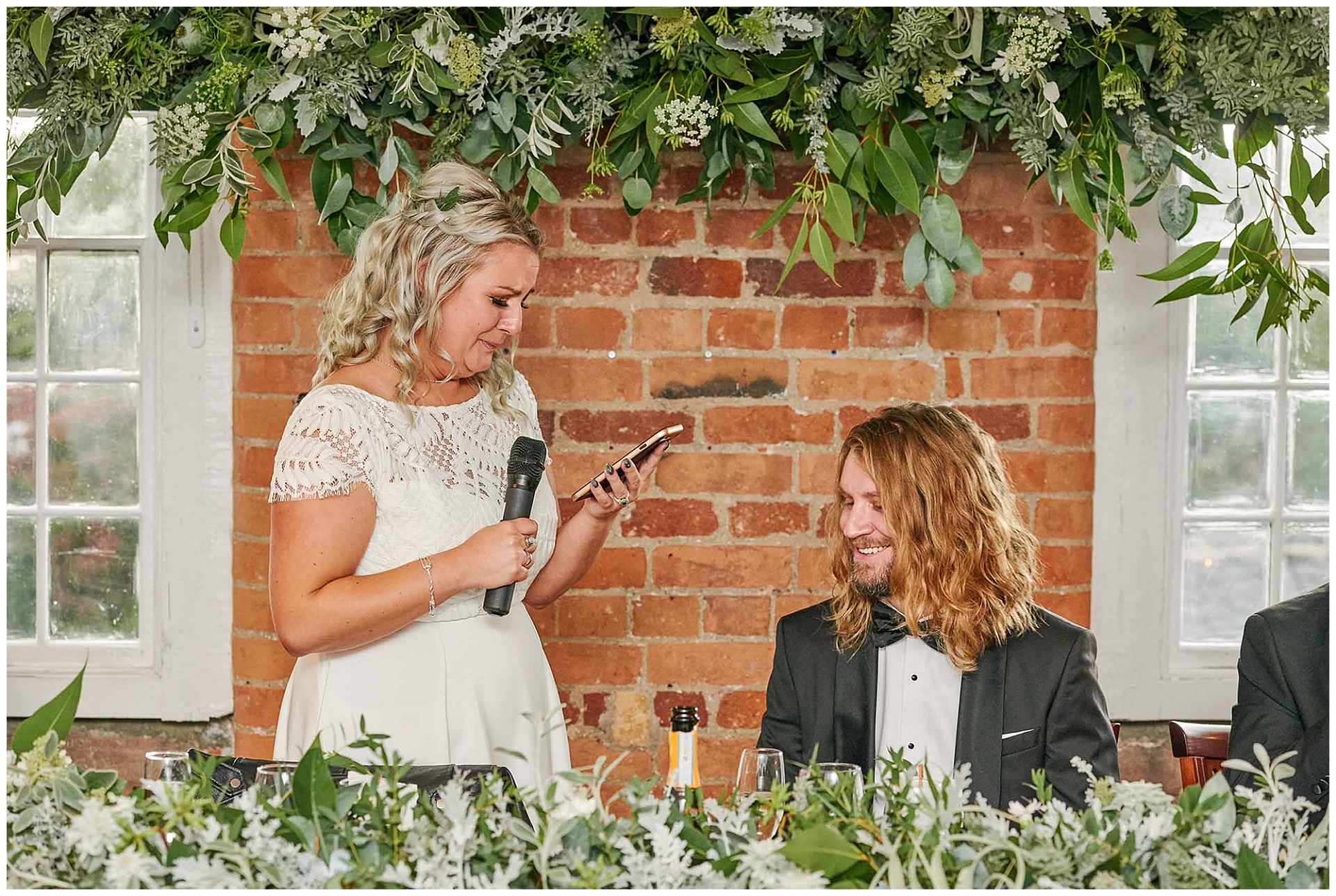 Bride crying whilst reading speech to the groom in front of a brick wall and green floral decorations at the West Mill in Derby.