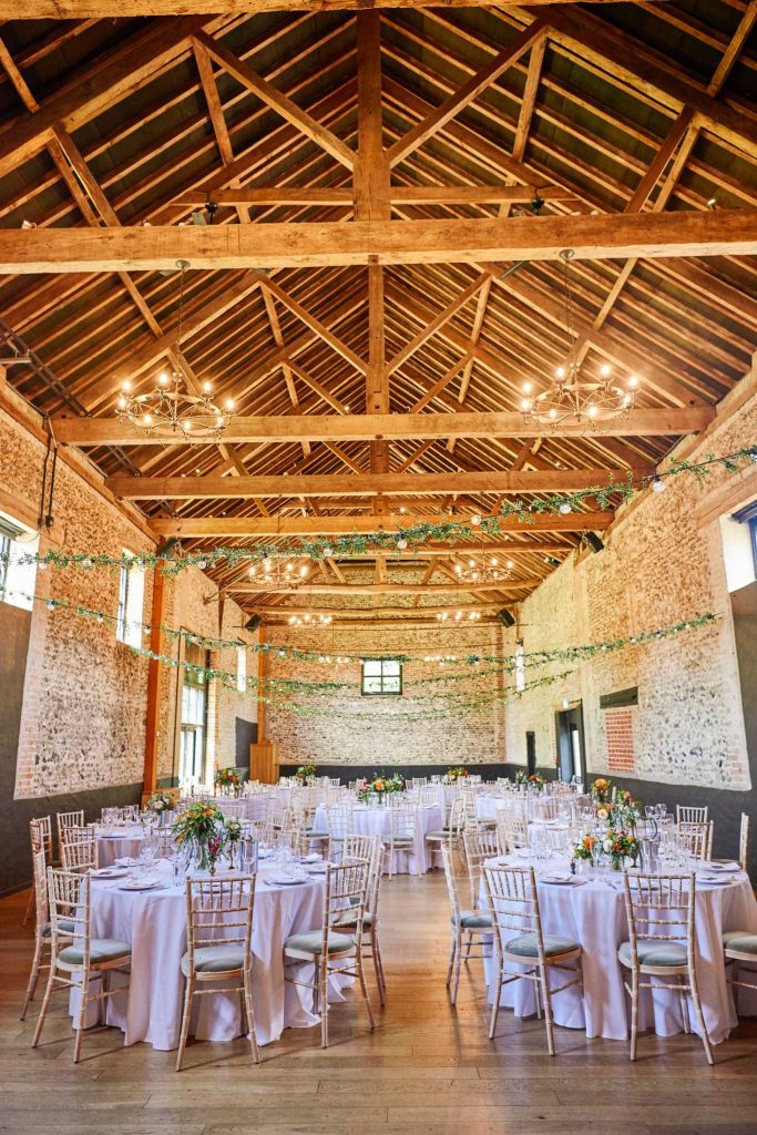 Empty wedding breakfast room with round tables, brick walls and rustic wooden beams at the Granary Estates in Cambridge.
