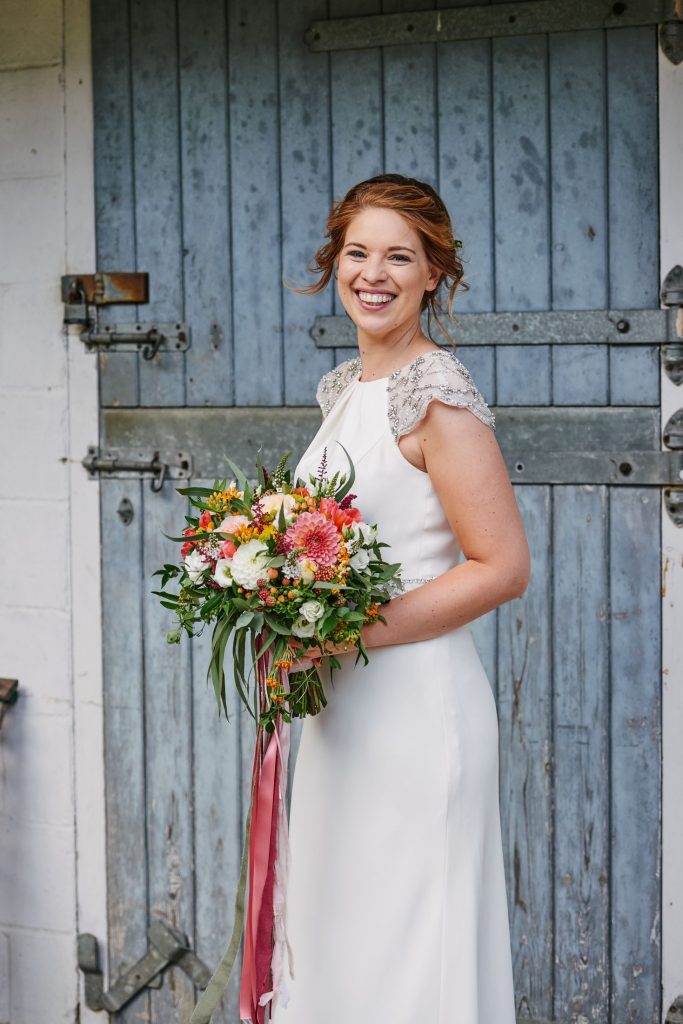 smiling bride holding colourful flowers and wearing a white wedding dress stood in front of a blue barn door in Cambridge