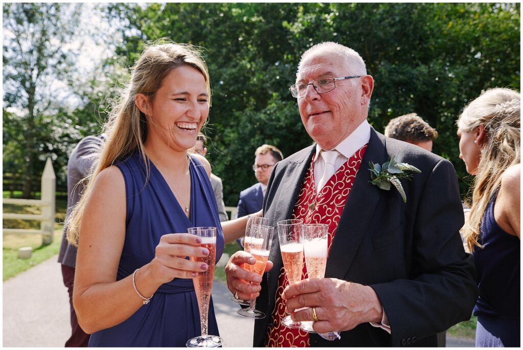 Wedding guests laughing whilst holding champagne and stood in the sunshine 
