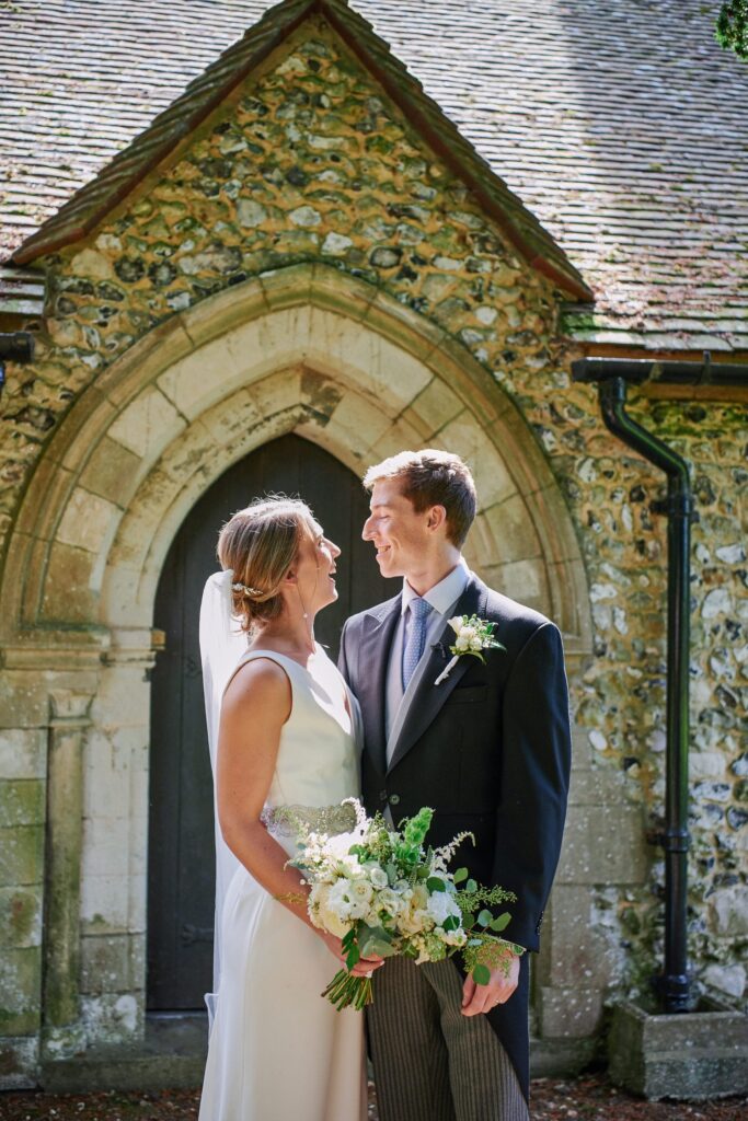 bride and groom smiling at each other whilst stood in the sunshine outside of church