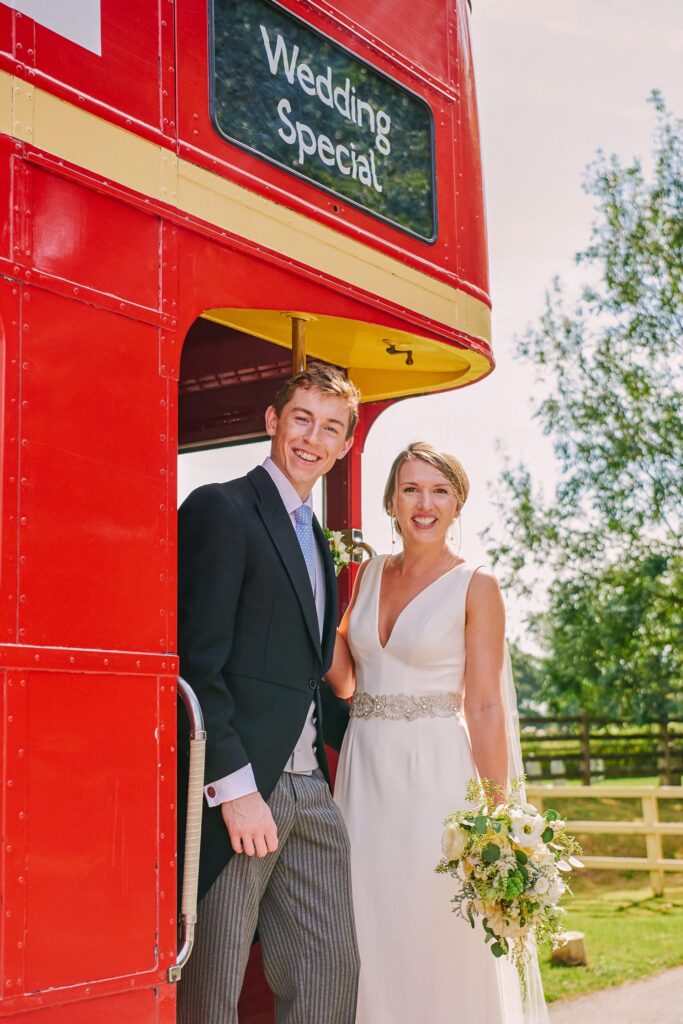 Bride and groom smiling on the back of a red London bus in the countryside 