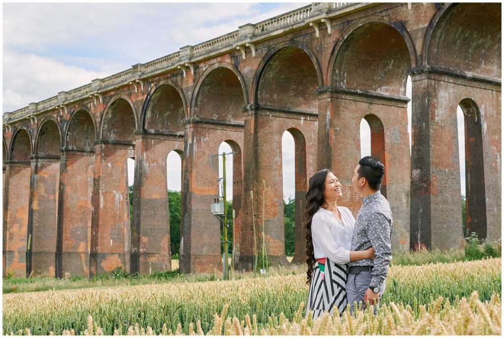 Engaged couple smiling at each other in a field in front of The Ouse Valley Viaduct