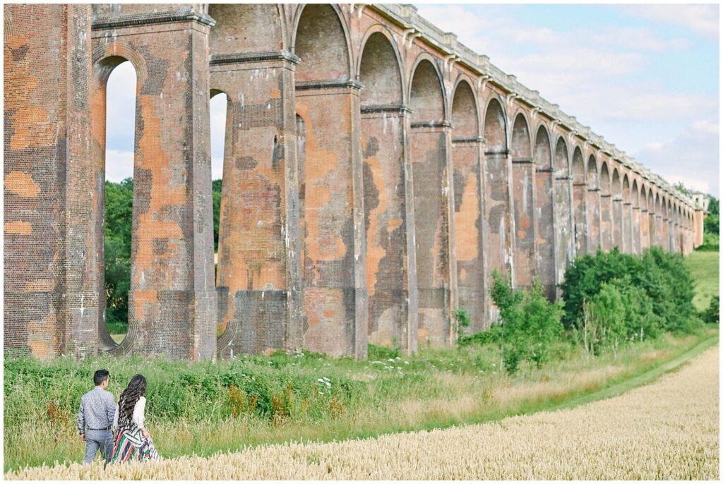 Engaged couple walking through a field in front of The Ouse Valley Viaduct train line