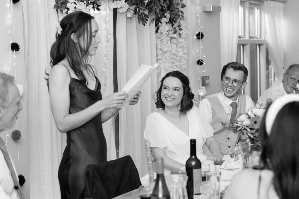 Bridesmaid giving speech while bride and groom smile during intimate wedding breakfast moment