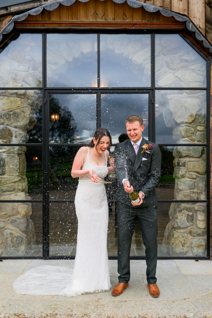 Bride and groom celebrating with champagne spray outside barn wedding venue