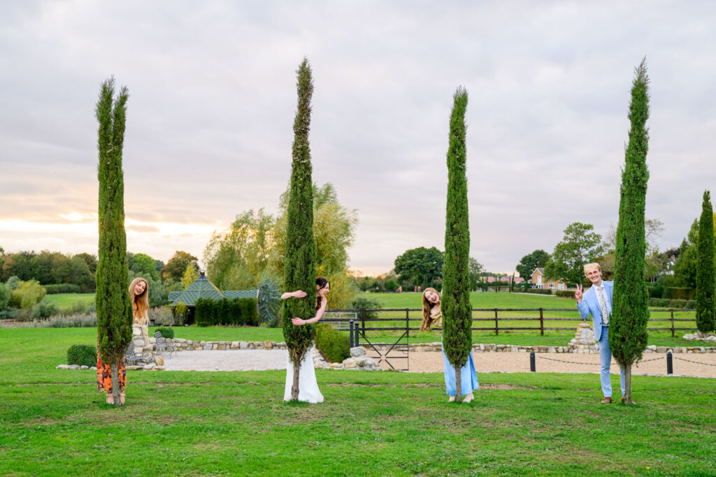 Wedding guests playing hide and seek behind trees during sunset reception at Willow Marsh Farm