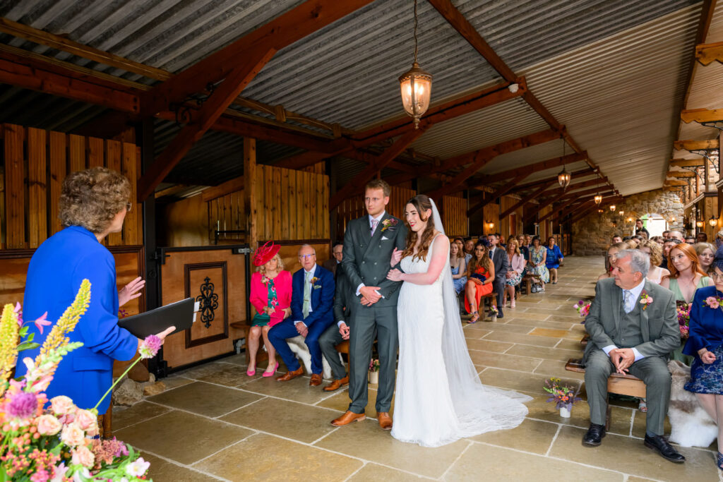Wedding ceremony taking place inside rustic barn venue with guests seated on wooden benches