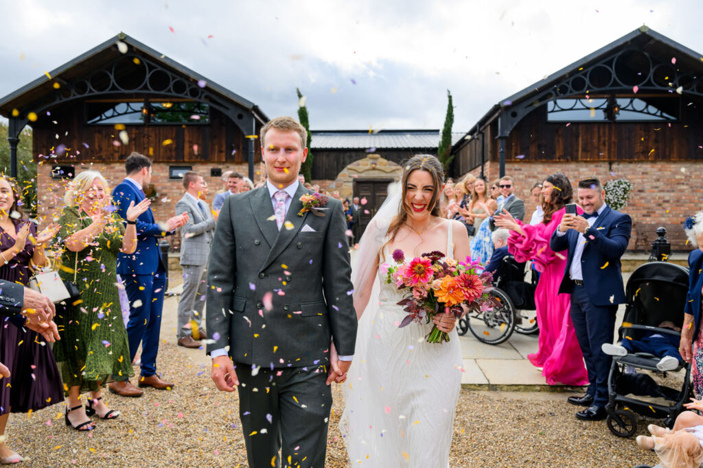 Bride and groom walking through colourful confetti outside Willow Marsh Farm barn