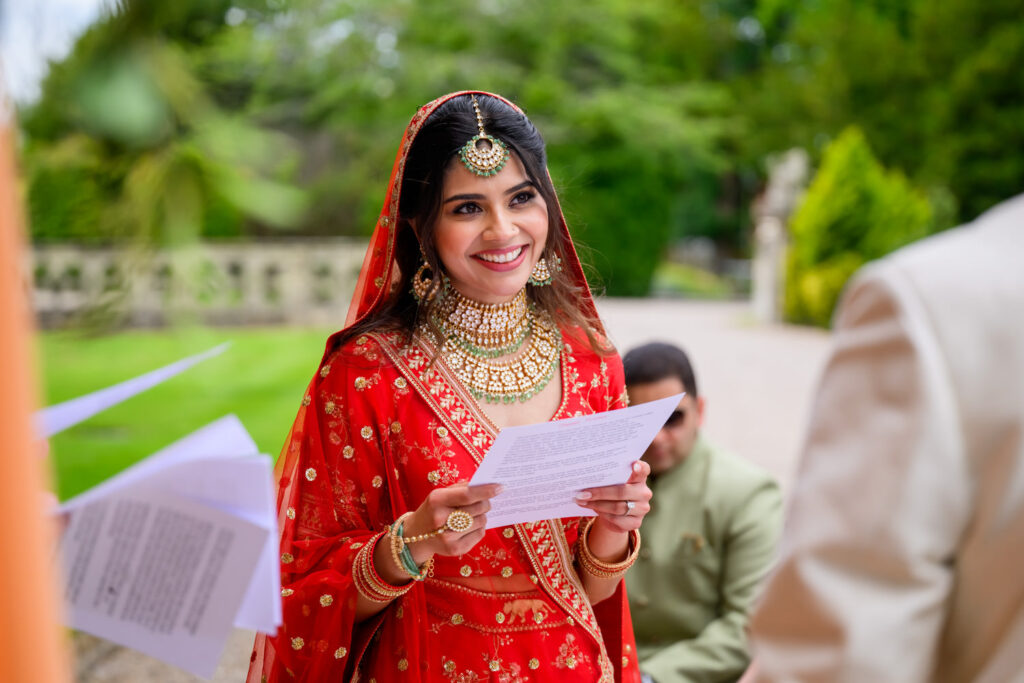 Bride wearing red and smiling as she reads her vows during the ceremony.