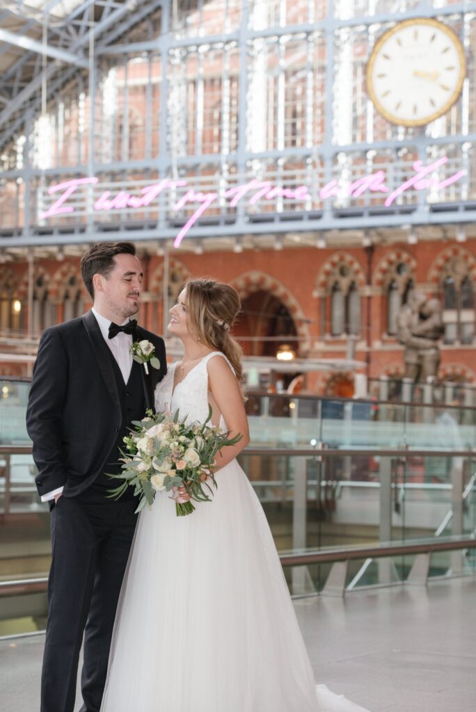 bride and groom smiling in front of the clock at St Pancras Eurostar station