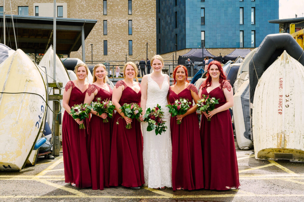 Bride stood with bridesmaids wearing red dresses in the boat yard