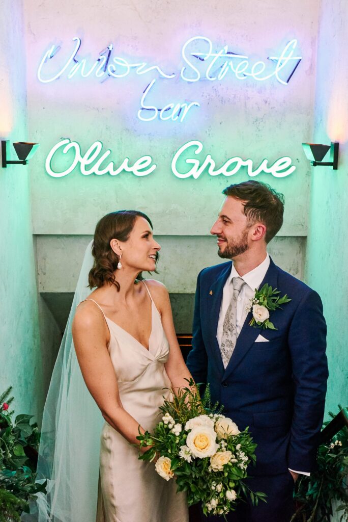 Wedding couple smiling in front of the neon Olive Grove sign at Union Street Café