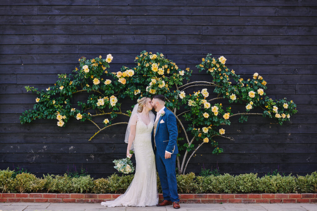 bride and groom kissing whilst stood in front of black barn & yellow roses 